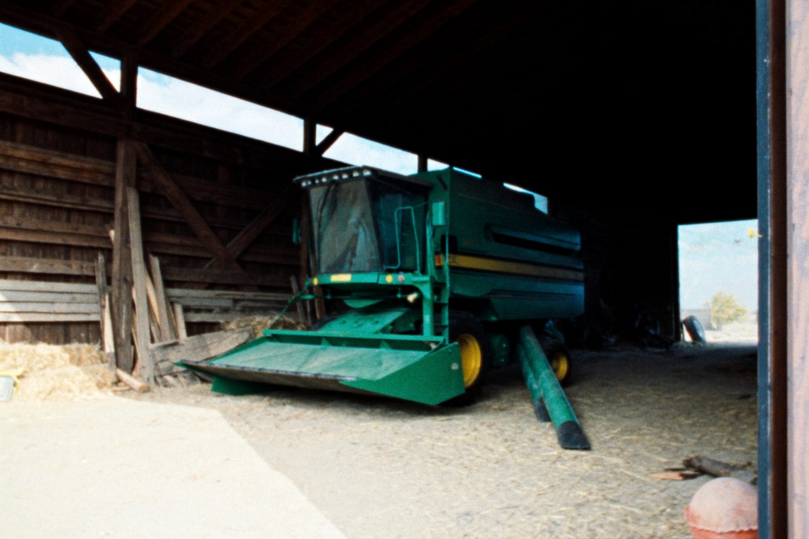 An older green combine harvester parked under the open bay of a pole machine shed, header detached and resting on the dirt floor beside it.