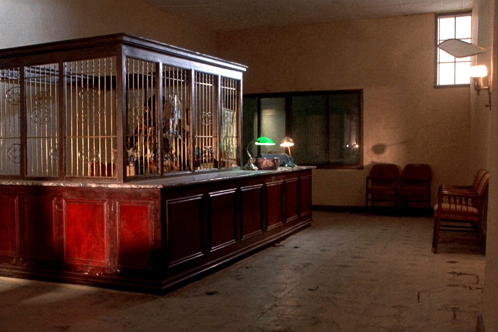 The inside of a small bank lobby late in the day, a wooden teller counter with the lamps switched on and empty chairs against the wall.
