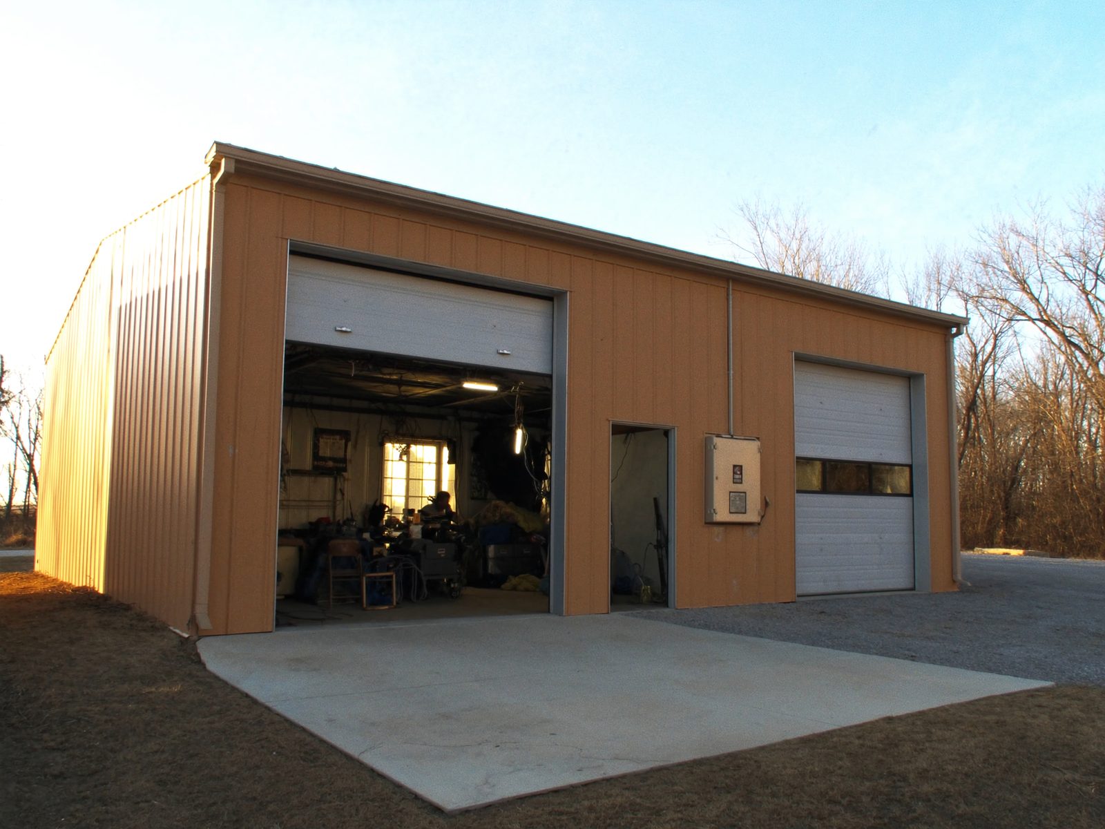 A tan steel-sided contractor shop building at low sun, one overhead door rolled up with work lights on inside, concrete apron and gravel drive.