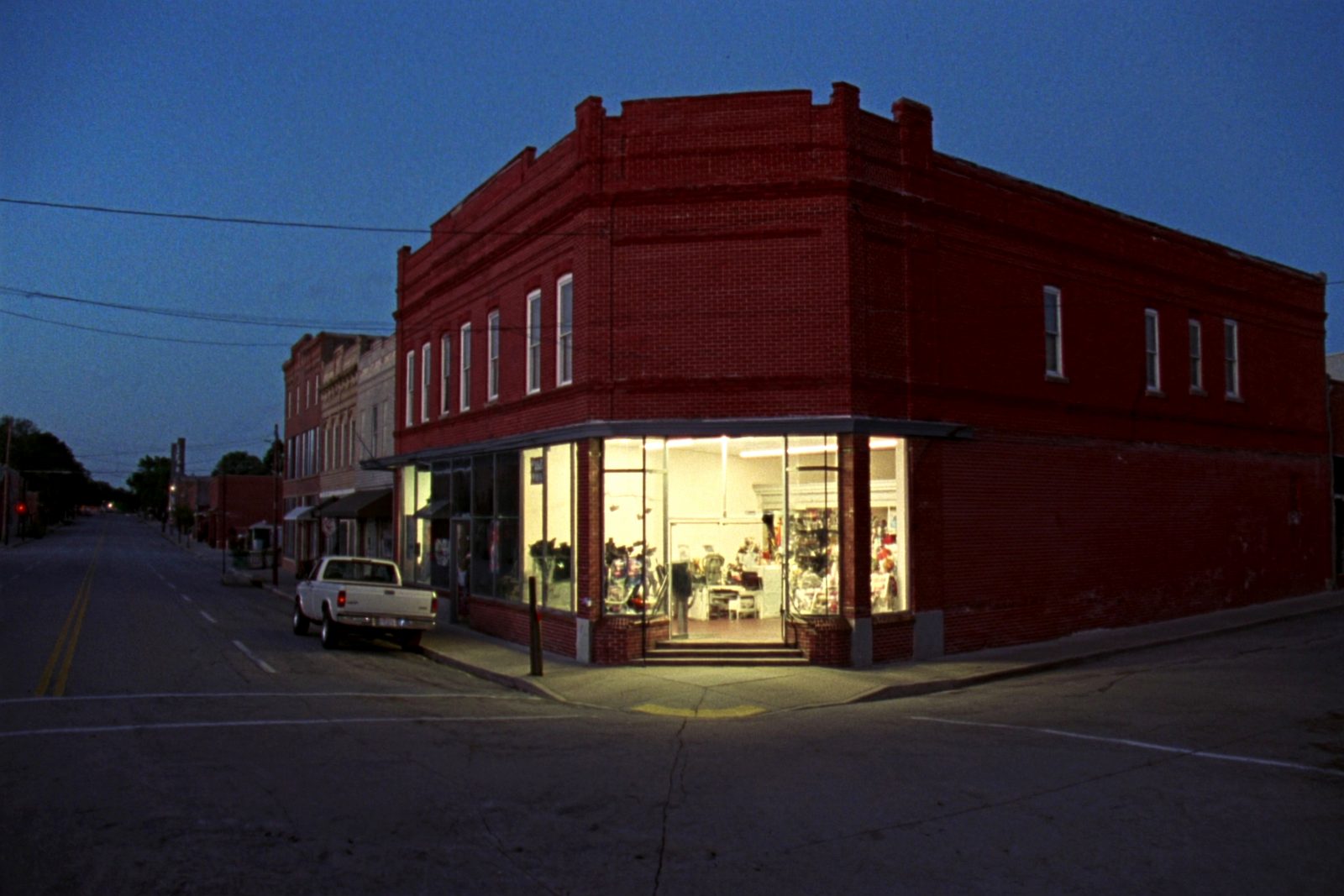 A plain two-storey red brick corner building on a small town square at dusk, warm lights on behind the ground floor glass, a pickup parked at the kerb.